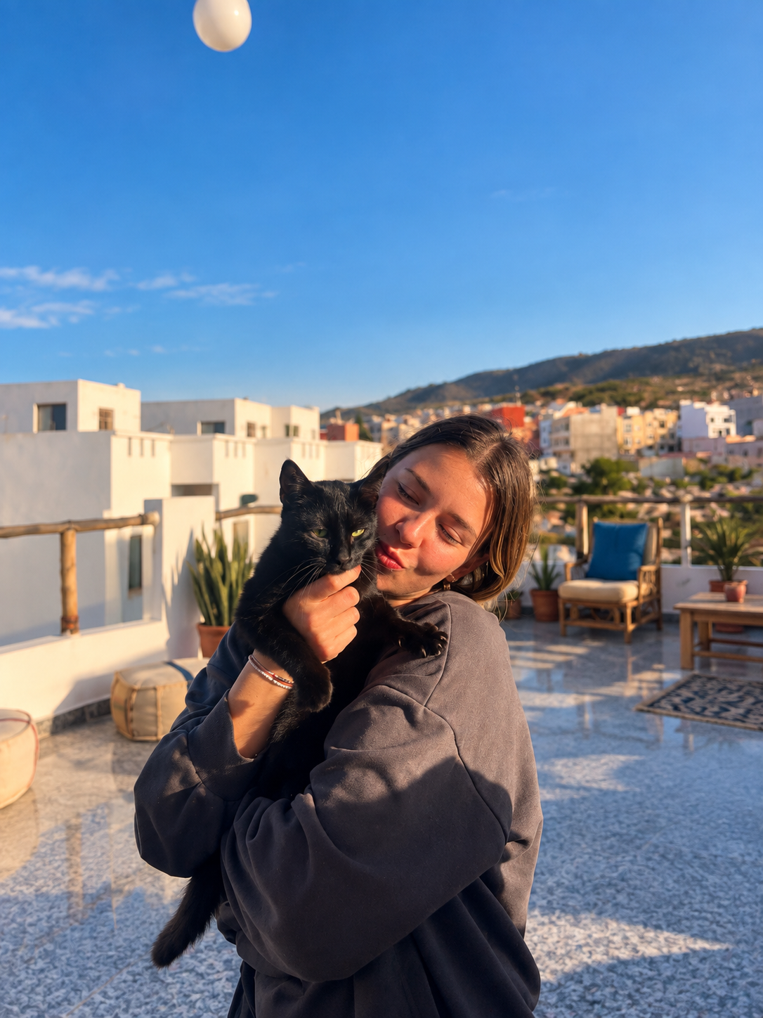 Voyageuse avec un chat noir sur un rooftop marocain à Taghazout, ciel bleu et maisons blanches